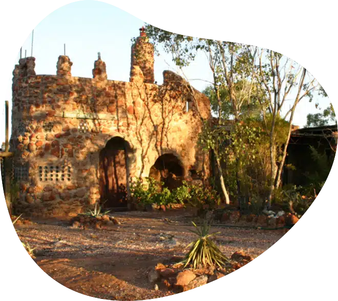 A rustic, castle-like stone building with arched doorways and small towers, surrounded by dry, rocky soil and desert plants under warm sunlight.