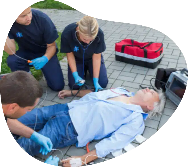 Three paramedics in navy uniforms attend to an unconscious man lying on a paved surface, with medical equipment and a red emergency bag beside them.