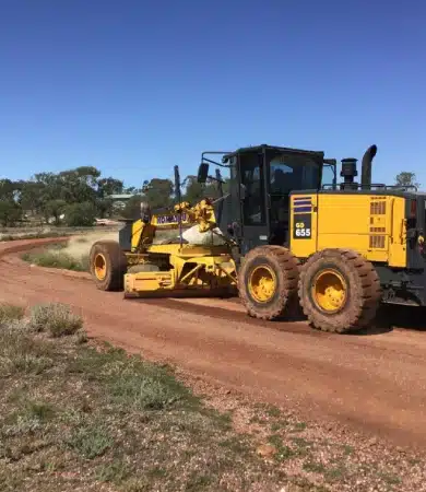 A yellow road grader drives along a curved dirt road in a rural area, with trees and grass visible under a clear blue sky.