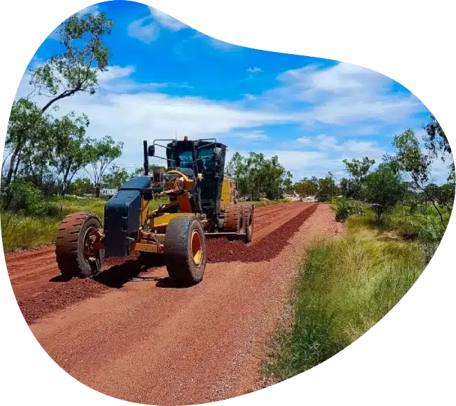 A yellow road grader levels a reddish gravel road in a rural area, surrounded by green vegetation and trees under a blue sky with scattered clouds.
