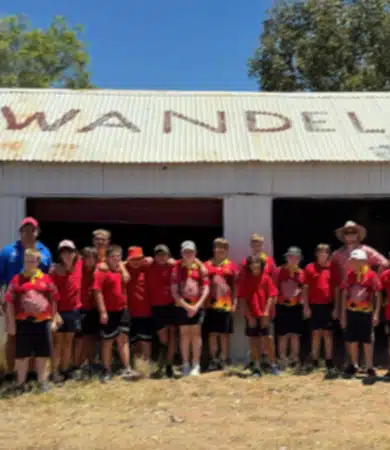 A group of children and a few adults wearing red shirts stand together in front of a corrugated metal building with "Wandel" partially visible on the roof. Trees and blue sky are in the background.
